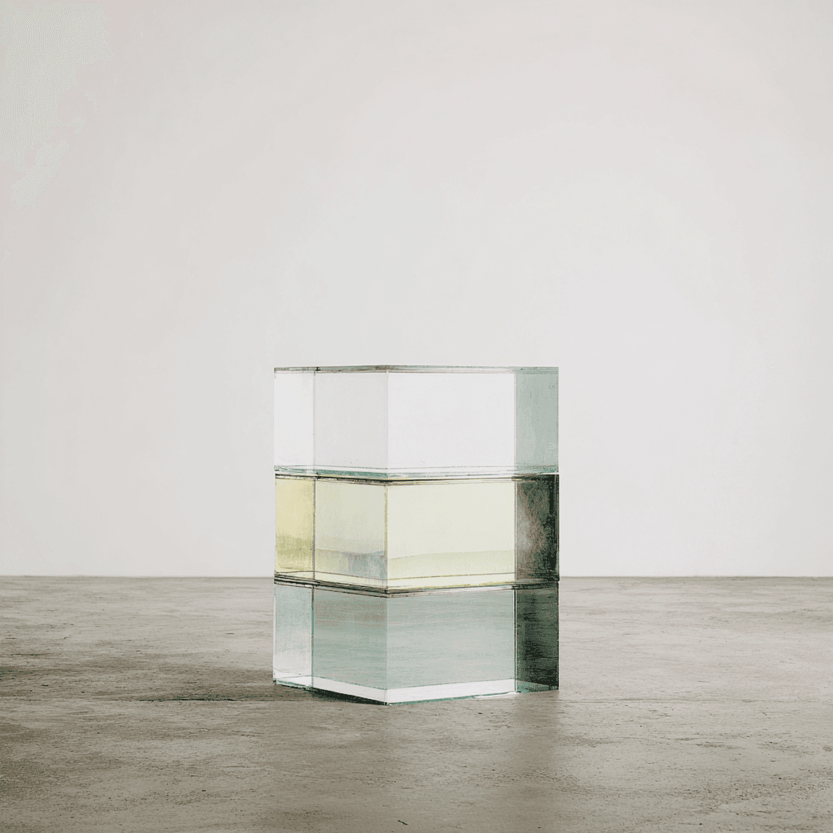 Stacked glass cube with layered translucent liquid on a concrete floor—minimal studio sculpture against a white wall.