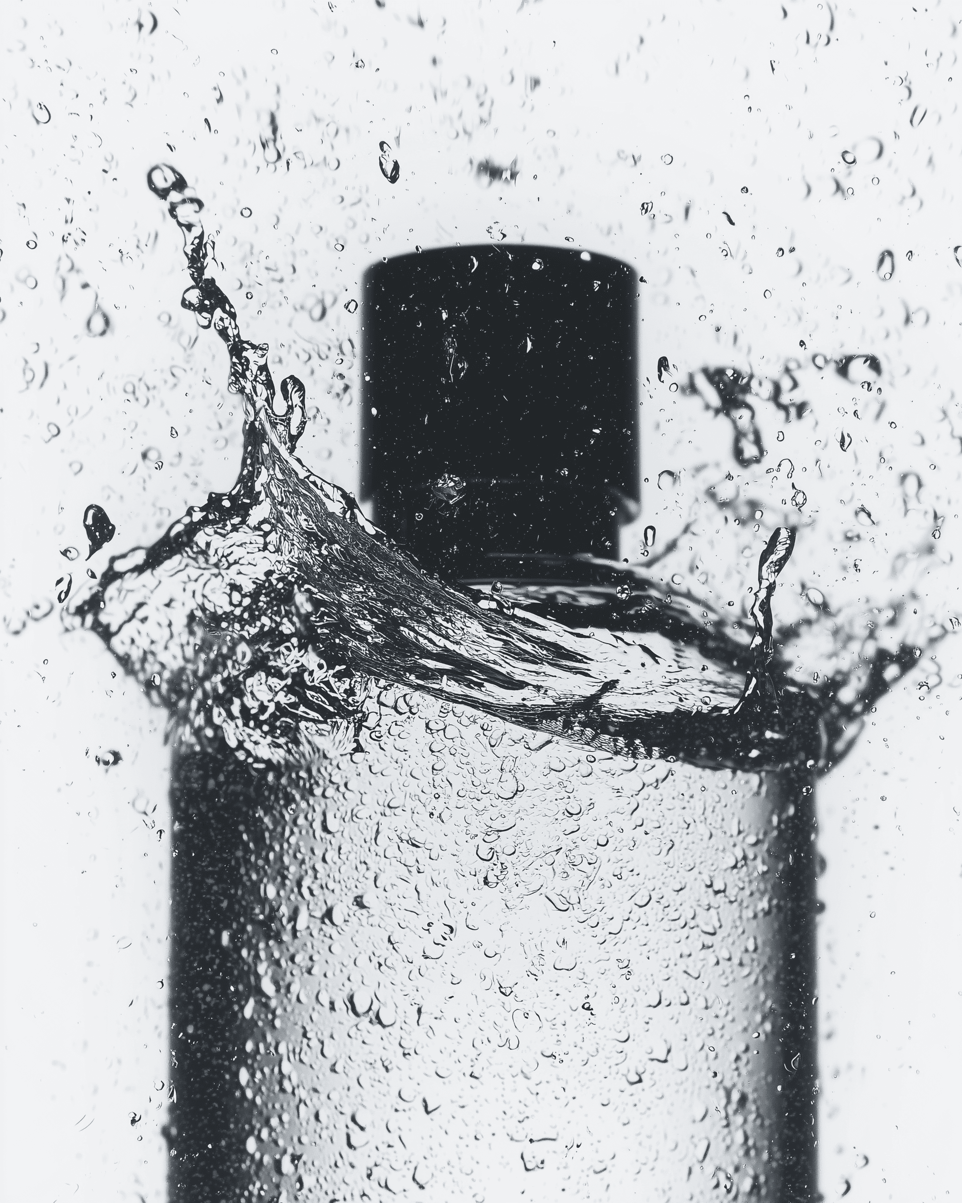 Monochrome product shot: black-capped cosmetic bottle with dynamic water splash on a clean white background.